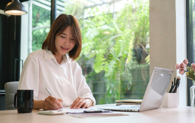 Smiling female office worker using laptop computer and making note on notebook.