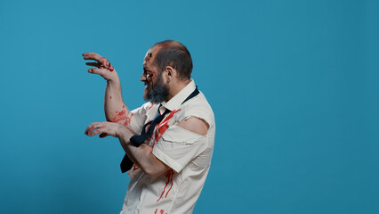 Scary looking brain-eating doing a funny dance on blue background. Spooky brain dead zombie with deep aned bloody wounds and scars dancing goofy while looking at camera. Studio shot - obrazy, fototapety, plakaty