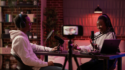 Obraz premium Content creator interviewing famous vlogger recording vlog using digital camera in home studio. African american couple broadcasting podcast using professional microphone and audio equipment.