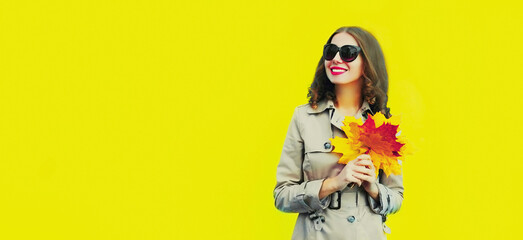 Portrait of beautiful young woman with yellow maple leaves wearing jacket, sunglasses on bright yellow background