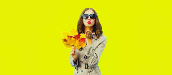 Portrait of beautiful young woman with yellow maple leaves blowing her lips sends kiss wearing jacket, sunglasses on bright yellow background