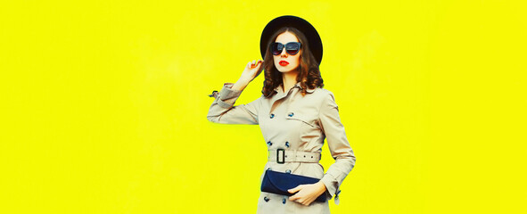 Portrait of stylish woman posing wearing black round hat, jacket and handbag clutch on bright yellow background