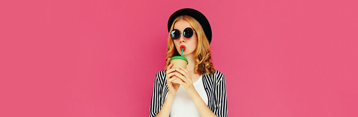 Portrait of stylish young woman drinking coffee or juice wearing striped black white shirt, round hat, sunglasses on pink background