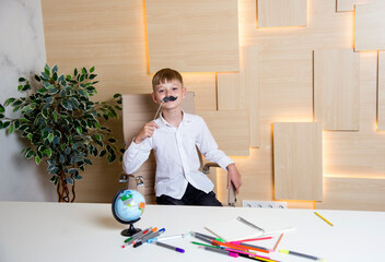 schoolboy in ukrainian embroidery with funny mustache sitting at the desk  at school with different colored penciles and globe - obrazy, fototapety, plakaty