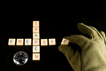 Eine Hand mit Handschuh legt den letzten Buchstaben zu den Wörtern Energie und sparen und ein Thermometer, das  18° C zeigt für eine niedrigere Raumtemperatur um Energie zu sparen.