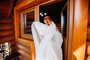 Cute red-haired bride in a satin white dress and with a white veil is posing on the balcony against...