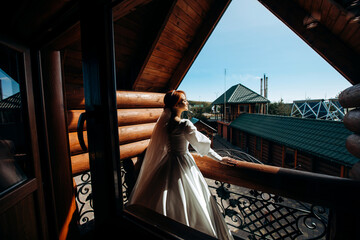 Pretty red-haired bride in a satin white dress is posing on the balcony on her wedding day.