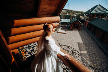 Luxury red-haired bride in a satin white dress is posing on the balcony on her wedding day.