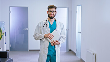 Good looking doctor man standing in front of the camera in the modern hospital corridor holding his...