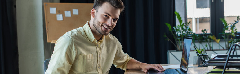Positive businessman in shirt using laptop and looking at camera in office, banner.