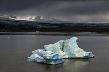 Fjallsarlon Glacier Lagoon, Iceland, on a stormy day. A blue iceberg floats in the lagoon and sunlight highlights the mountains behind.