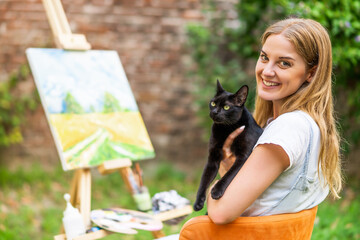 Woman enjoys   spending time with her cat and painting  on canvas outdoor.