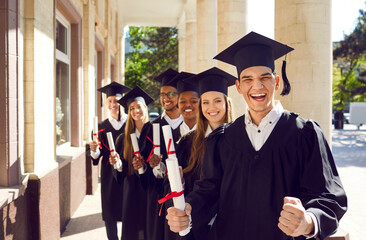Happy excited university graduates rejoice in successful completion of their studies. Students in gowns and academic caps with diplomas in hands posing on near walls and columns of university.