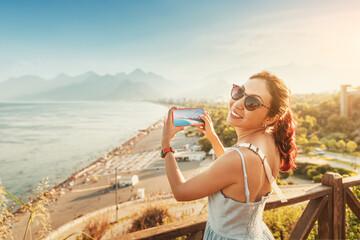 Naklejka premium Travel blogger girl takes high-quality and vivid photos on the camera of his new expensive smartphone of the famous Konyaalti beach from a scenic viewpoint in Antalya, Turkey