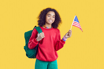 Smiling African American teen girl isolated on yellow studio background hold America flag in hands. Happy biracial female student with backpack study abroad. International education.