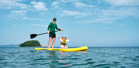 A father rides his little daughter in a life jacket on a big yellow SUP board in the sea. Active...