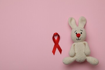 Cute knitted toy bunny and red ribbon on pink background, flat lay with space for text. AIDS disease awareness
