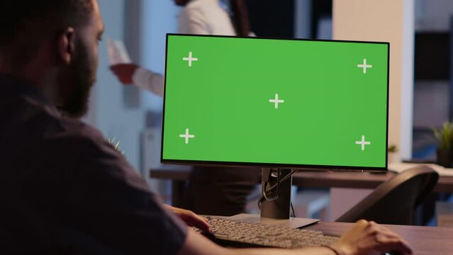 African american man looking at computer with greenscreen, analyzing isolated display on desktop monitor. Using blank copyspace template with mockup screen and chroma key background.