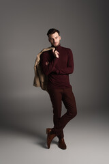 Full length of stylish man in turtleneck holding jacket and looking at camera on grey background.