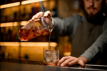 Hand of bartender holds mixing cup with strainer and poured drink into glass