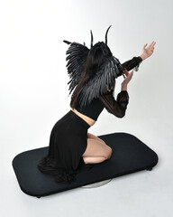 full length portrait of beautiful asian model with dark hair, wearing black gothic skirt costume, angel feather wings with horned headdress. Kneeling  pose  isolated on studio background.