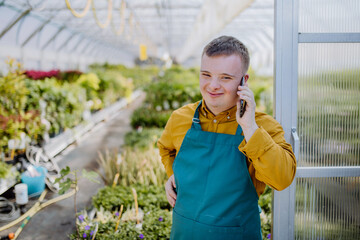 Young employee with Down syndrome working in garden centre, standing in door of greenhouse and...