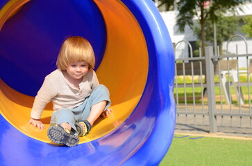 Fototapeta na wymiar A little boy rides down a slide on a playground near the house. Yard in a modern neighborhood. The kid rejoices in summer and a sunny day. Concept: kindergarten, child care, behavior, entertainment, a