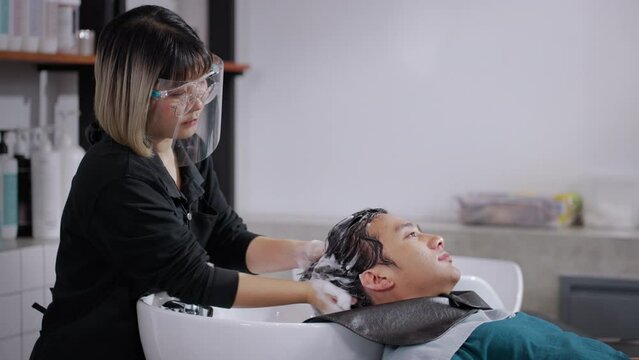 Asian attractive young male get hair washed by hairdresser in salon. Professional hair stylist woman give service and hair wash and massage to handsome man customer lying down on salon washing bed.