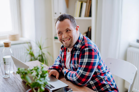 Young Man With Down Syndrome Sitting At Desk In Office And Using Laptop, Looking At Camera And Smiling.