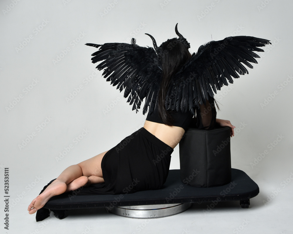 Wall mural full length portrait of beautiful asian model with dark hair, wearing black gothic skirt costume, angel feather wings with horned headdress. kneeling  pose  isolated on studio background. - Wall murals