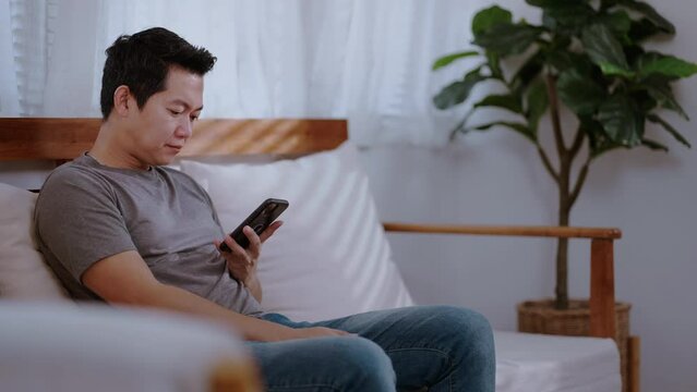 Asian handsome man read online news on mobile phone in morning at home. Happy attractive male sit on sofa in living room enjoy holiday weekend, using smartphone communication with technology in house.