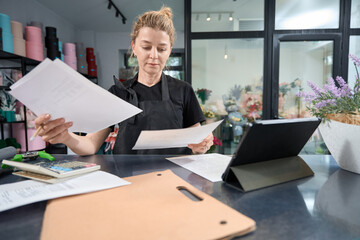 Focused flower shop owner sitting at counter looking through paperwork
