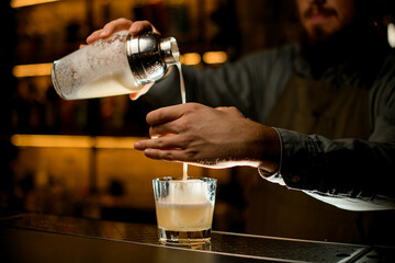 barman holds shaker and pours cold cocktail into glass filtering it through sieve