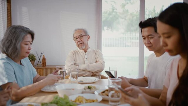Asian family using phone while eating food not attention to senior man. Upset Elderly grandfather feel unhappy and angry from Ignorance son, daughter and kid at home. Family and social issue concept.