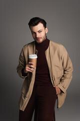 Stylish man in brown jacket holding coffee in paper cup isolated on grey.