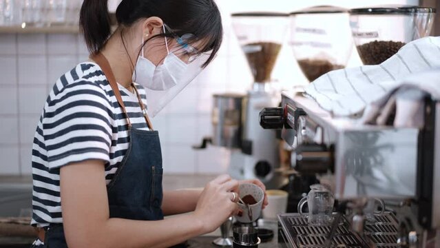 Asian waitress cafe woman wear face mask making coffee for customer. Attractive young girl specialist barista using grinder machine make espresso or latte coffee on slow bar in coffeeshop restaurant.