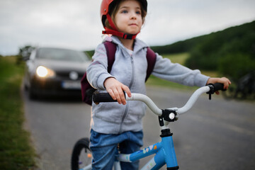 Portrait of excited little girl riding bike on road with car behind her, road safety education concept.