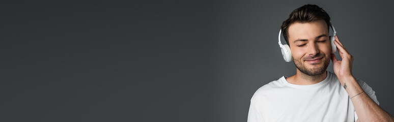 Bearded man smiling while listening music in headphones isolated on grey, banner.