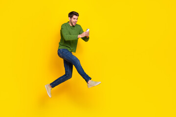 Fototapeta na wymiar Photo of funky excited guy wear green sweater jumping writing modern device empty space isolated yellow color background