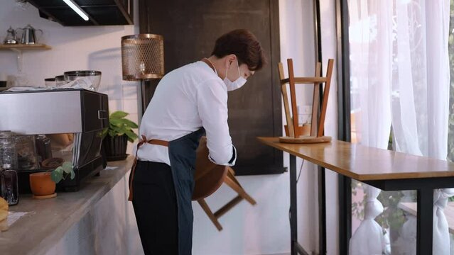 Asian cafe owner put chair on floor to clean and arranging furniture. Young waiter or cafeteria staff wear protective mask and cleaning coffee shop before open due to covid-19 pandemic in restaurant.