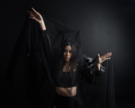 portrait of beautiful asian model with dark hair, wearing black gothic skirt costume, angel feather wings with horned headdress. Posing with gestural hands  on dark silhouette  studio background.