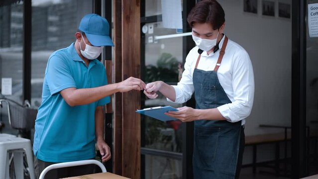 Asian delivery man wear mask receive box product from restaurant worker. Handsome business male waitress giving take away food box to postman deliver food at coffee shop, New normal lifestyle concept.