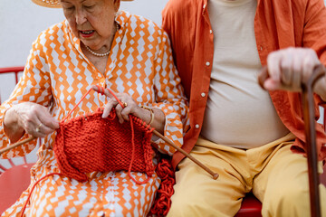 Senior woman sitting in the garden on the bench with his husband and knitting red scarf.
