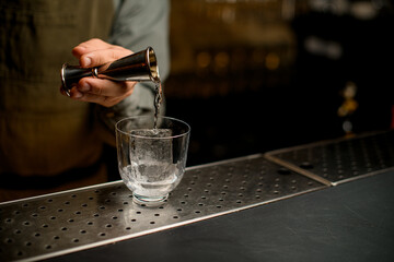 hand of bartender holds jigger and pours drink into transparent glass with ice