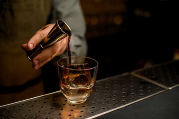 Close-up view of hand of bartender holding steel jigger and pours drink into transparent glass with an ice cube on bar