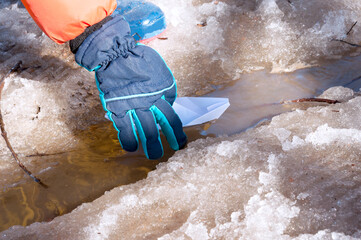 Kid's hand in winter gloves puts white paper boat to a spring stream among the snowy shores