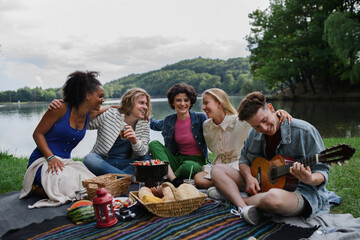 Group of friends having fun on picnic near a lake, sitting on blanket eating and playing guitar.