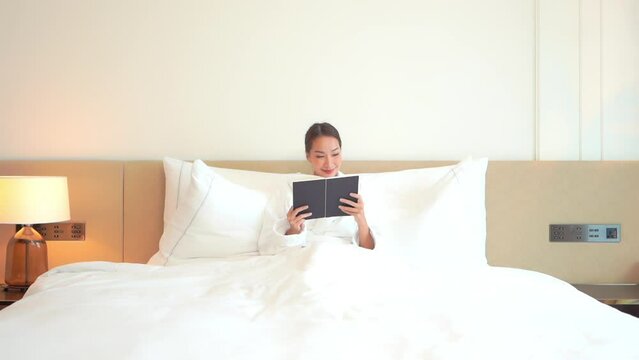 A pretty woman enjoys the perfect vacation morning and relaxes with a good book while sitting up in bed. 