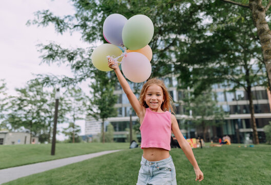 Little blond girl posing with baloons in city and looking at camera.