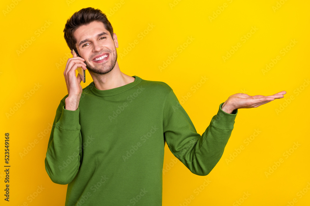 Wall mural photo of cute funny young guy wear green sweater talking modern device holding arm empty space isola